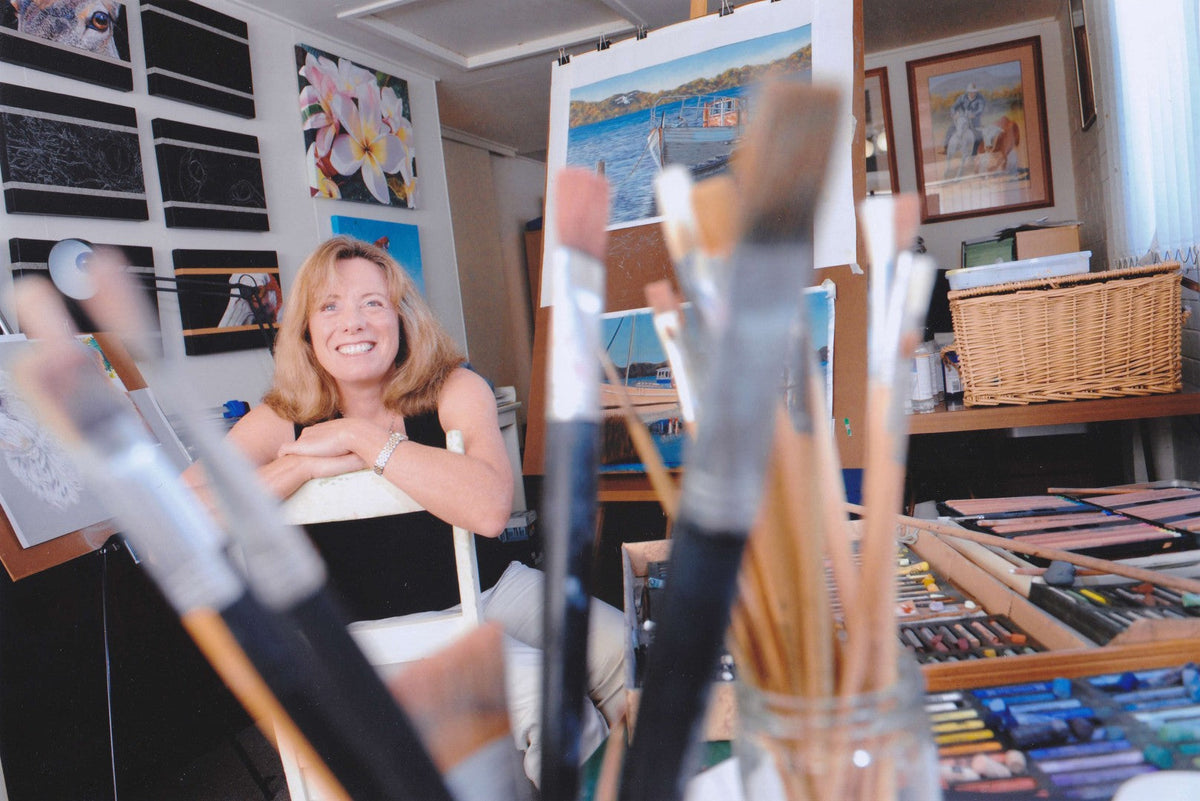 Sally McLean happy in her studio surrounded by artworks and creative art supplies such as brushes paints and pastels.